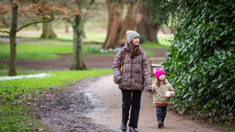 An adult and child walk in the Lacock woodland. They are both wearing winter hats and coats and are looking at the trees.